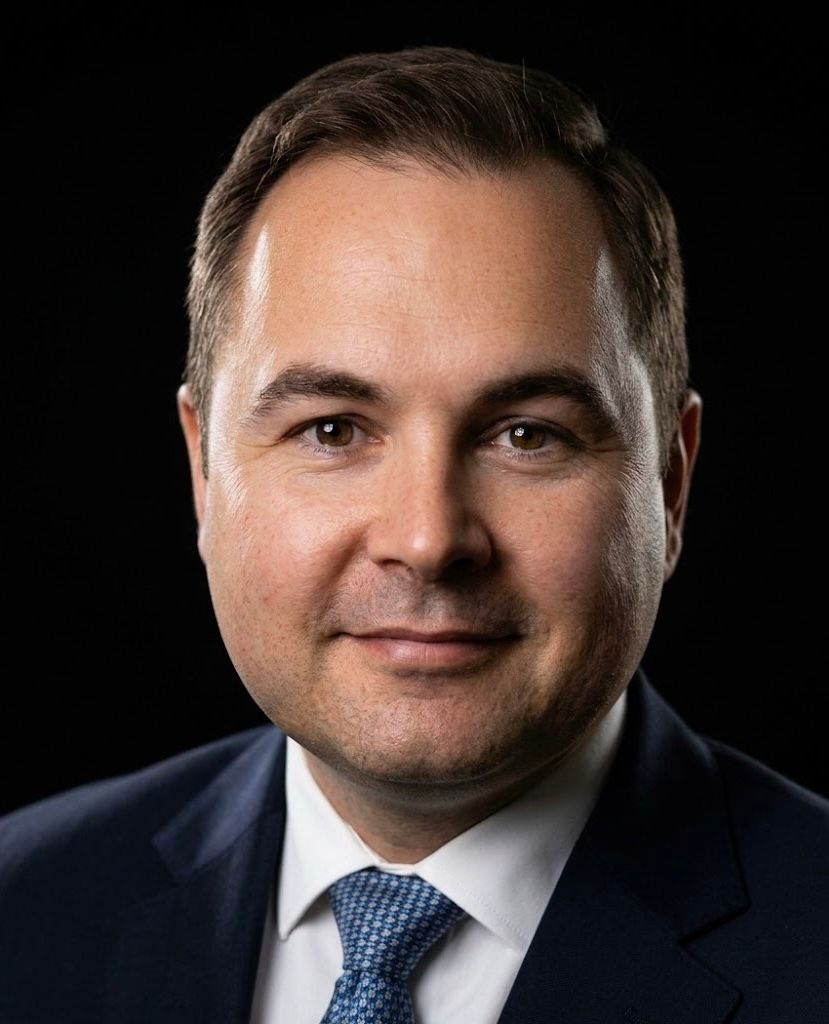 Studio portrait of a man in a dark suit and blue tie against a black background