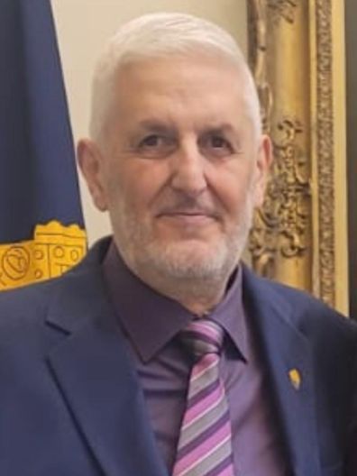 A man in a suit and tie stands in front of a flag