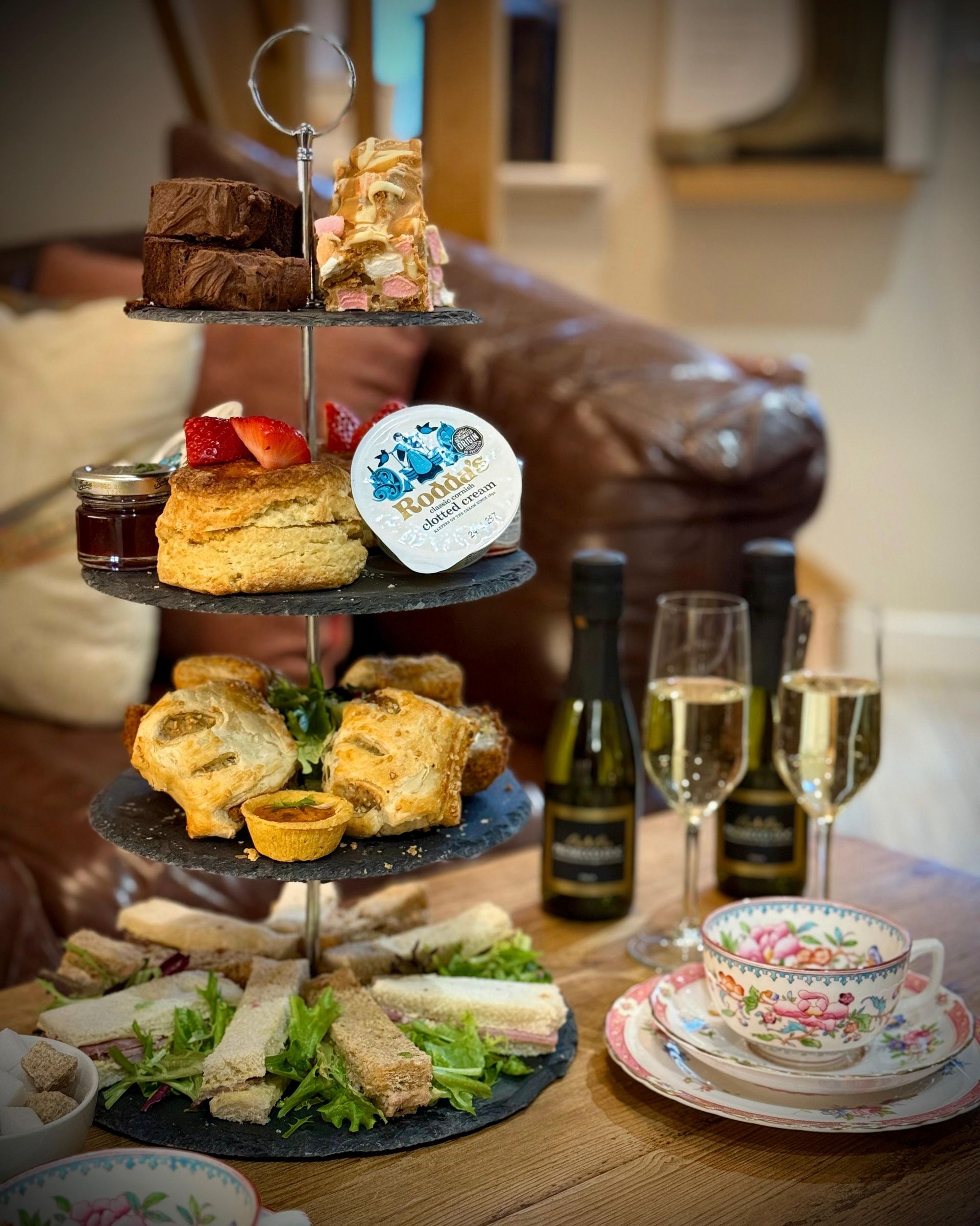Afternoon tea set: sandwiches, pastries, scones with jam, cake, and champagne, in a cozy setting.