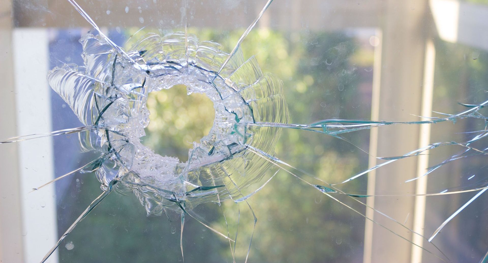 Broken window pane with a circular impact point and radiating cracks, overlooking trees.