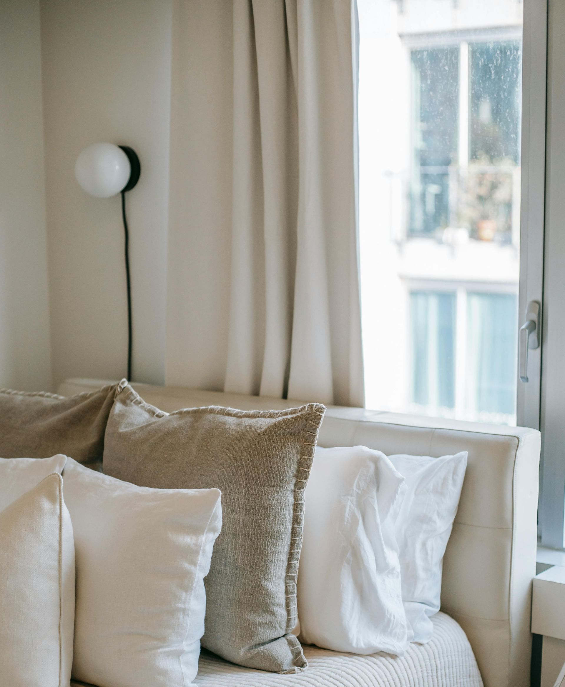 Bed with beige pillows and white curtains near a window, a black lamp is mounted on the wall.