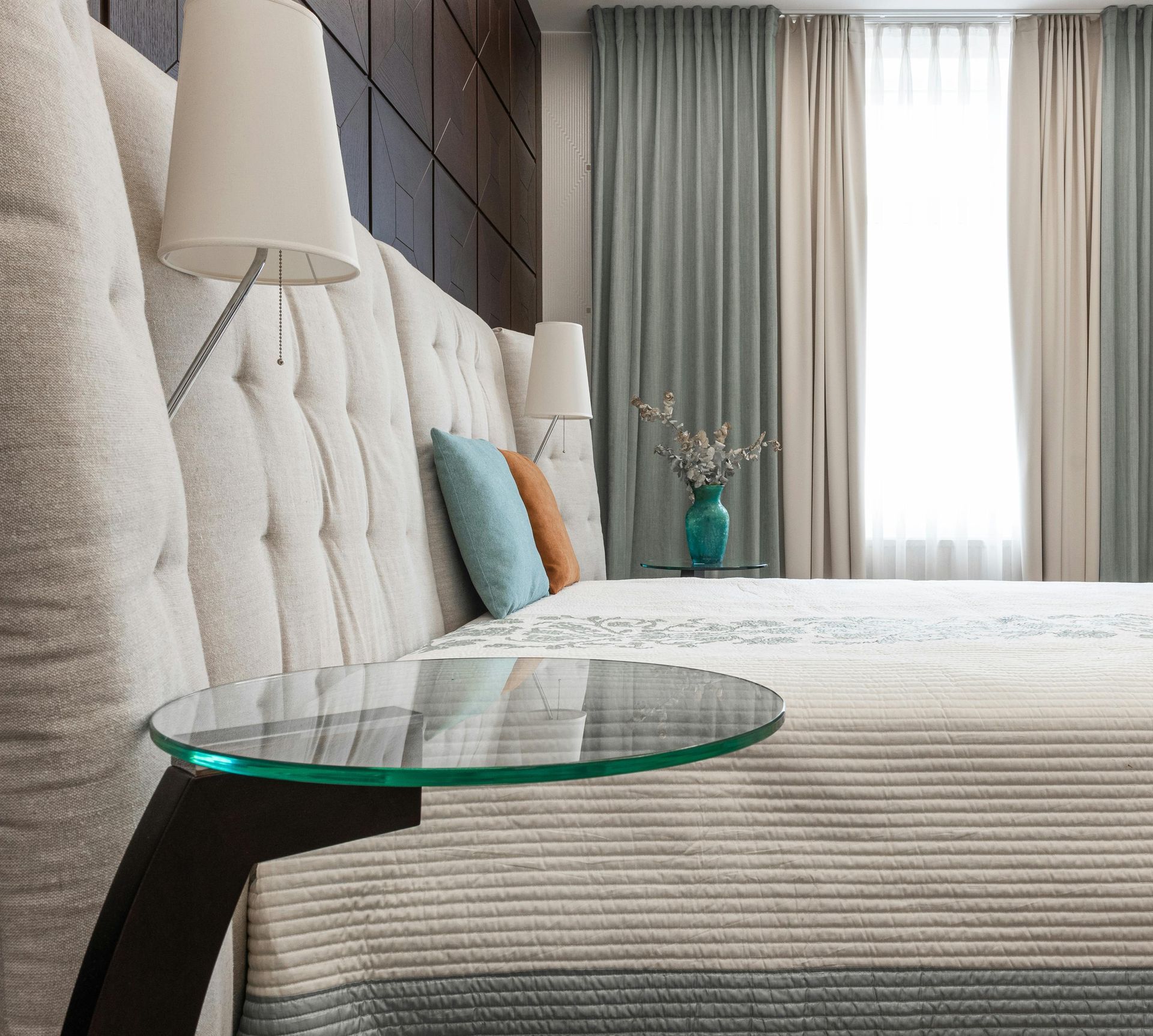 Bedroom with a large tufted headboard, a glass side table, and light blue curtains.
