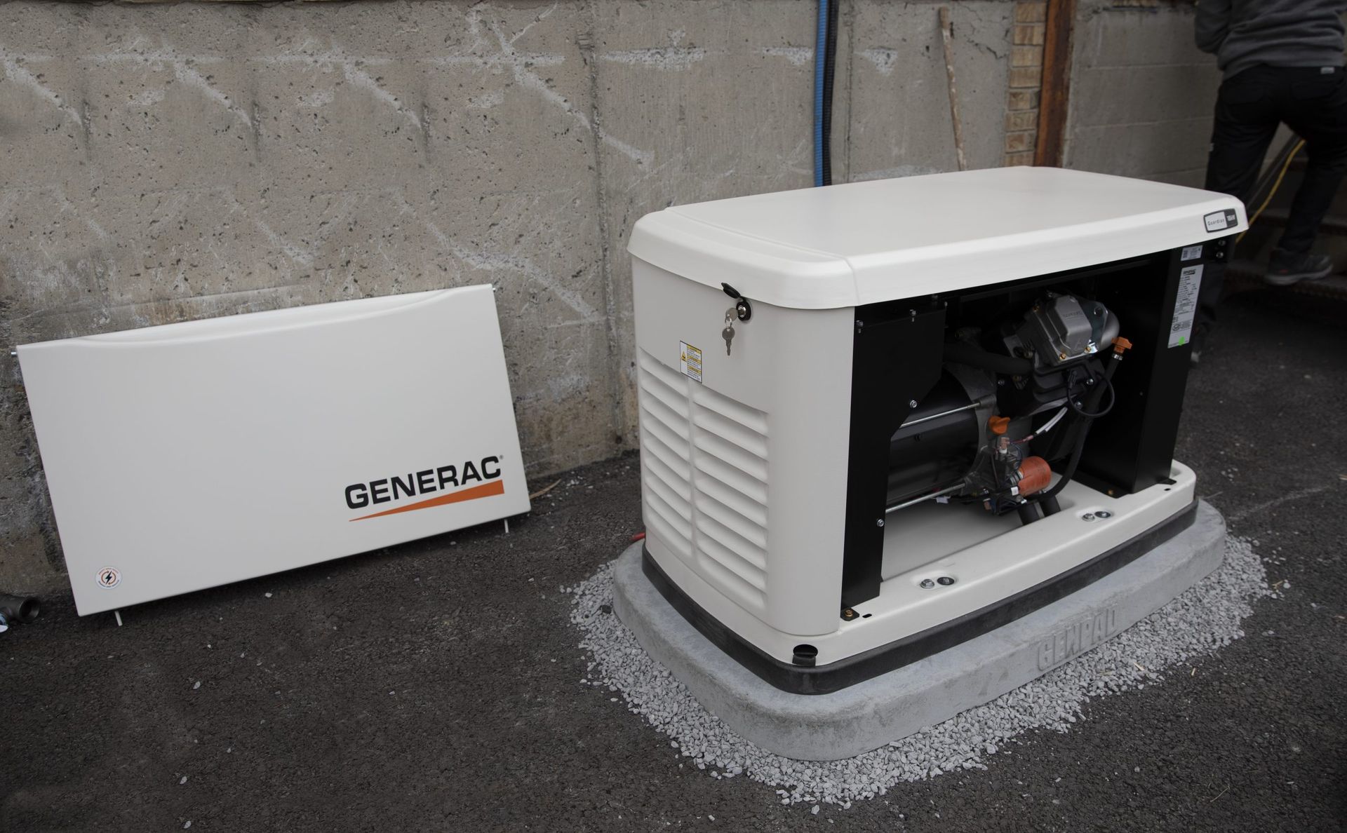 Un générateur domestique Generac sur une base de gravier, avec son panneau latéral retiré, près d'un mur en béton.