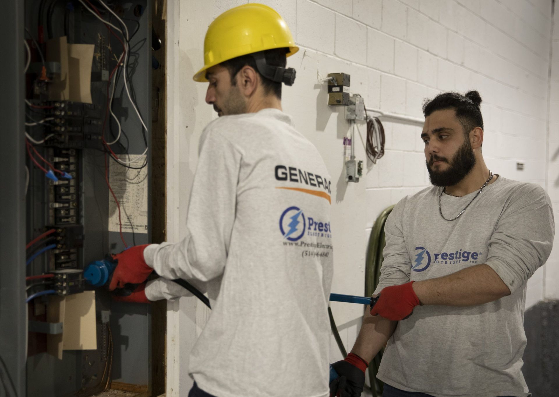 Two electricians working on an electrical panel. One wears a hardhat. Indoors, white walls.