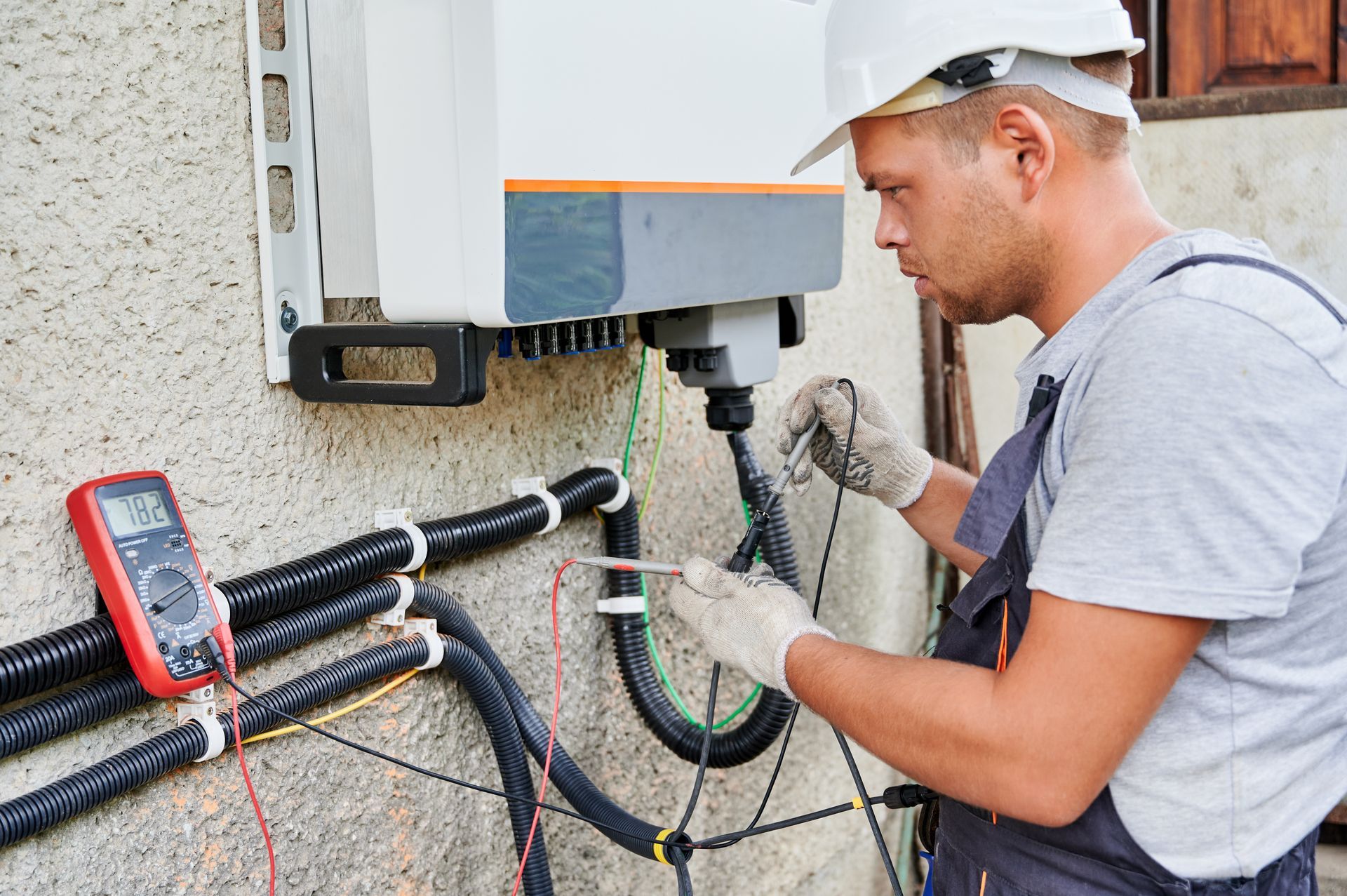 Electrician testing wires on a wall-mounted solar inverter with a multimeter; outdoors.