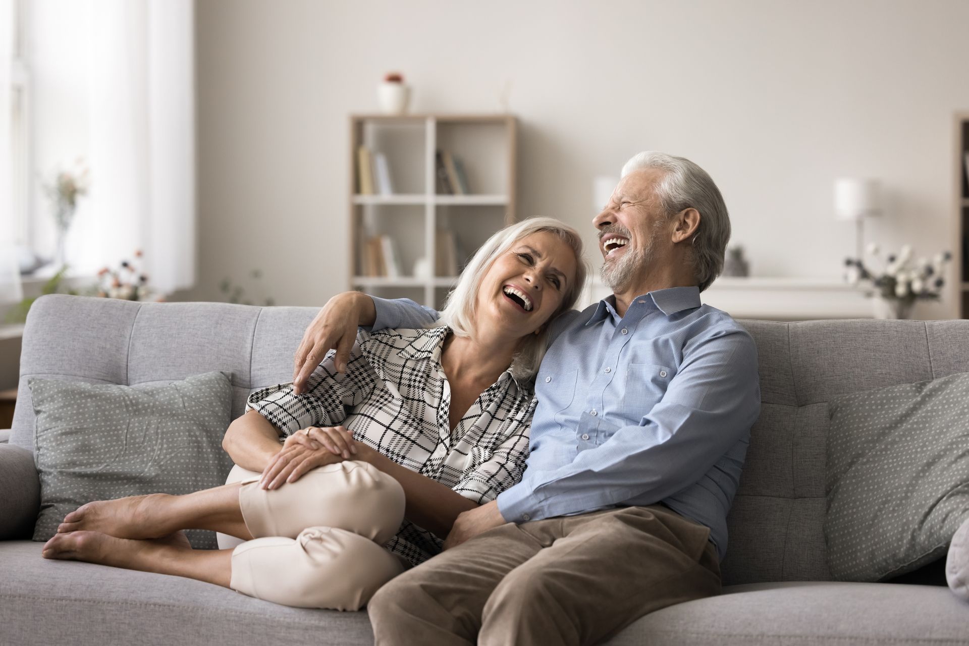 Elderly couple laughing together on a gray couch at home; man's arm around woman.