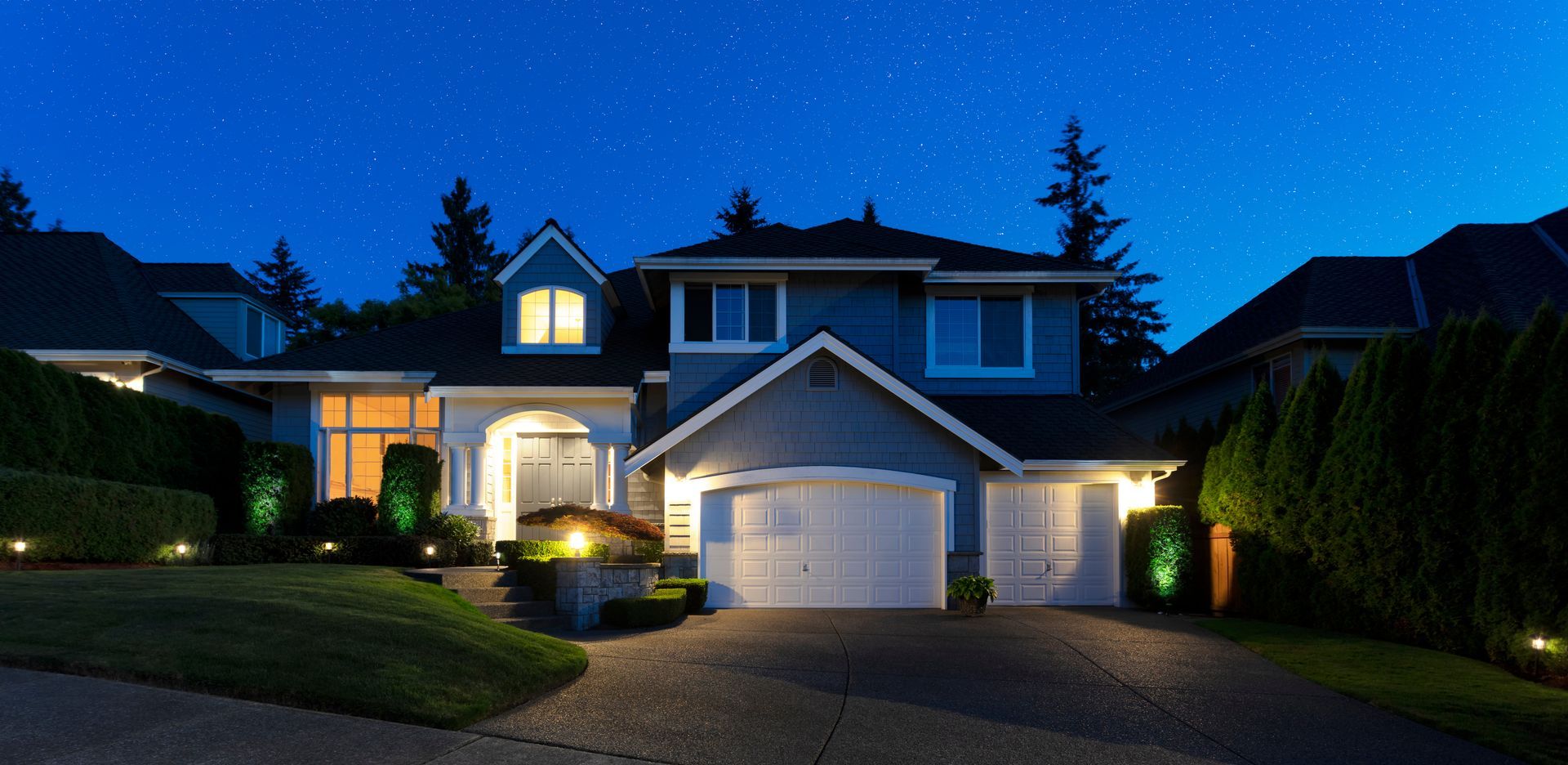 Une maison bleue avec des portes de garage blanches et des lumières illuminant la pelouse avant sous un ciel étoilé.