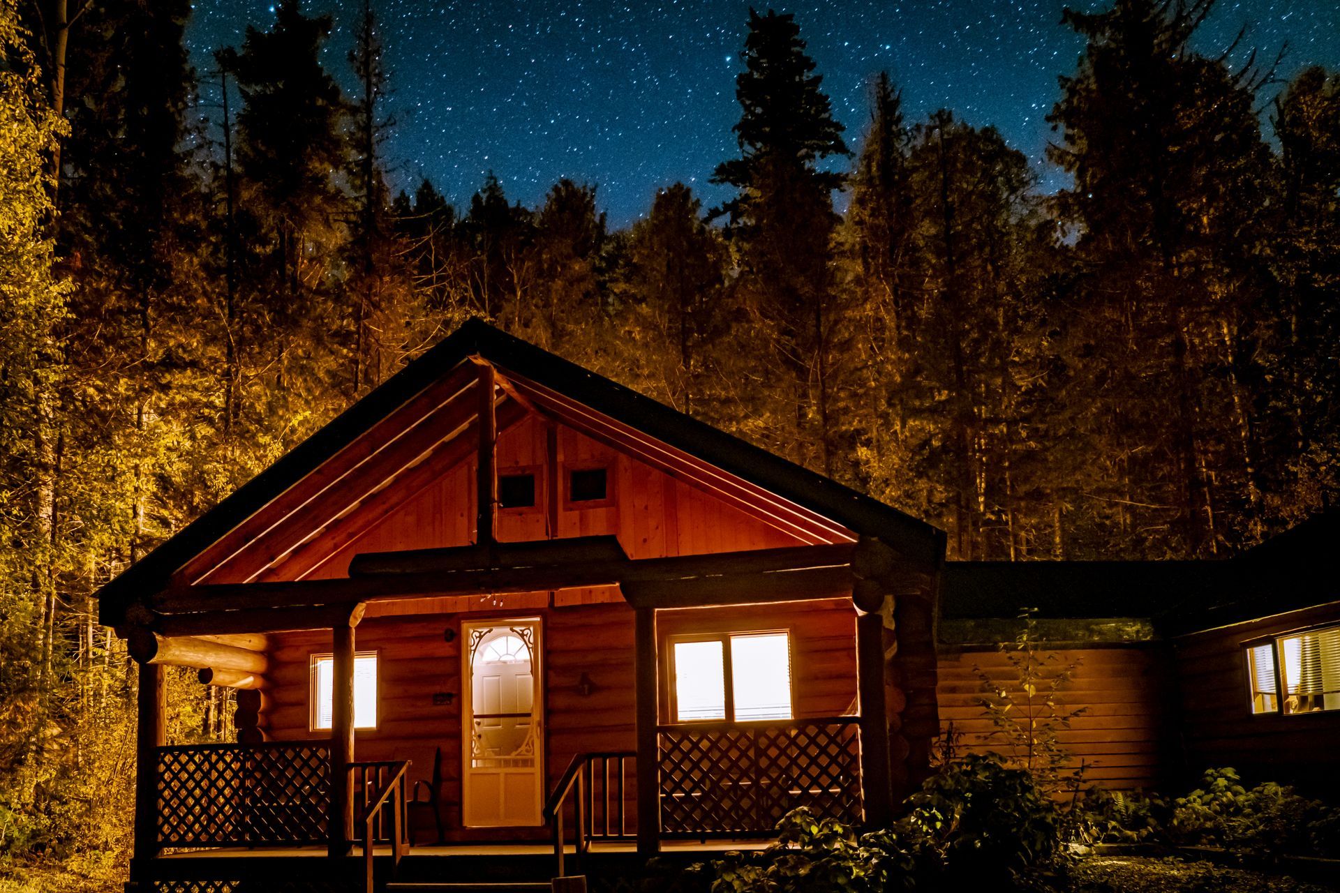 Cabane en bois la nuit, éclairée de l'intérieur, entourée d'arbres sombres sous un ciel étoilé.