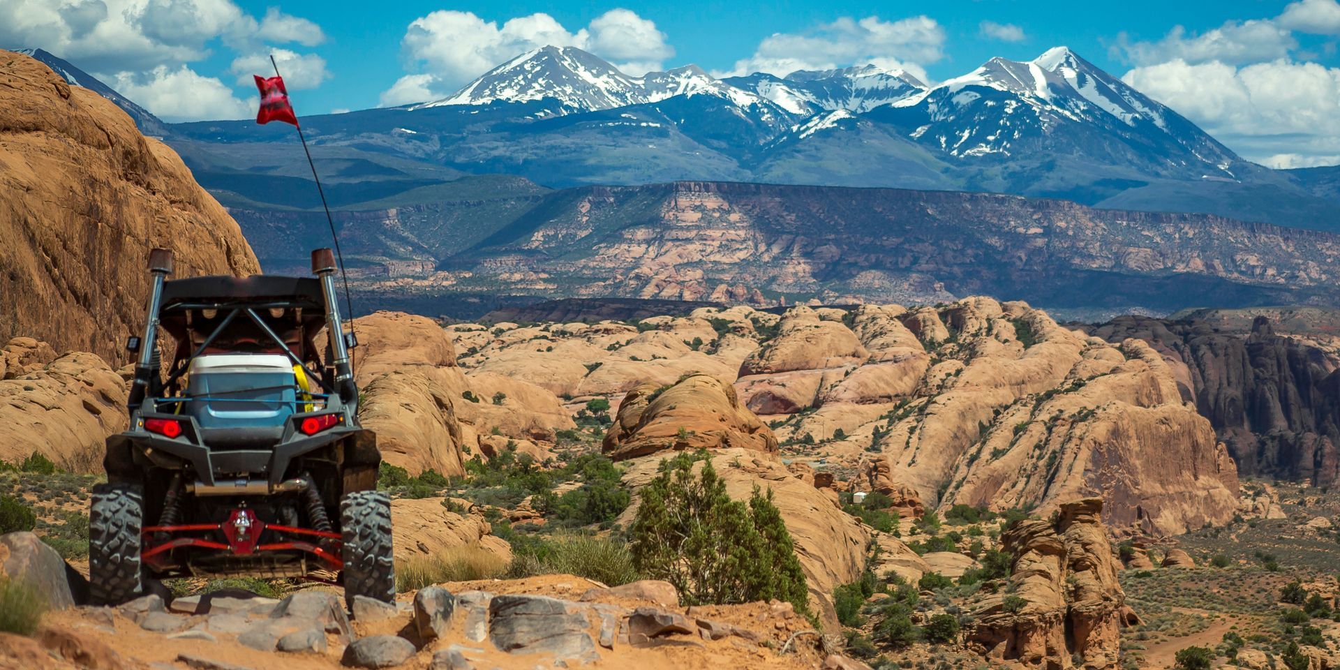 A dark-colored UTV sits on a rocky ridge, overlooking a vast desert canyon landscape with snow-capped mountains beyond.