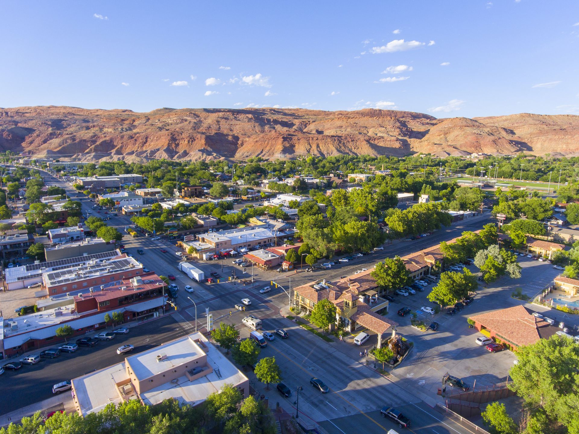 Aerial view of Moab, Utah, featuring desert cliffs overlooking a town with green trees and roads under a clear blue sky.