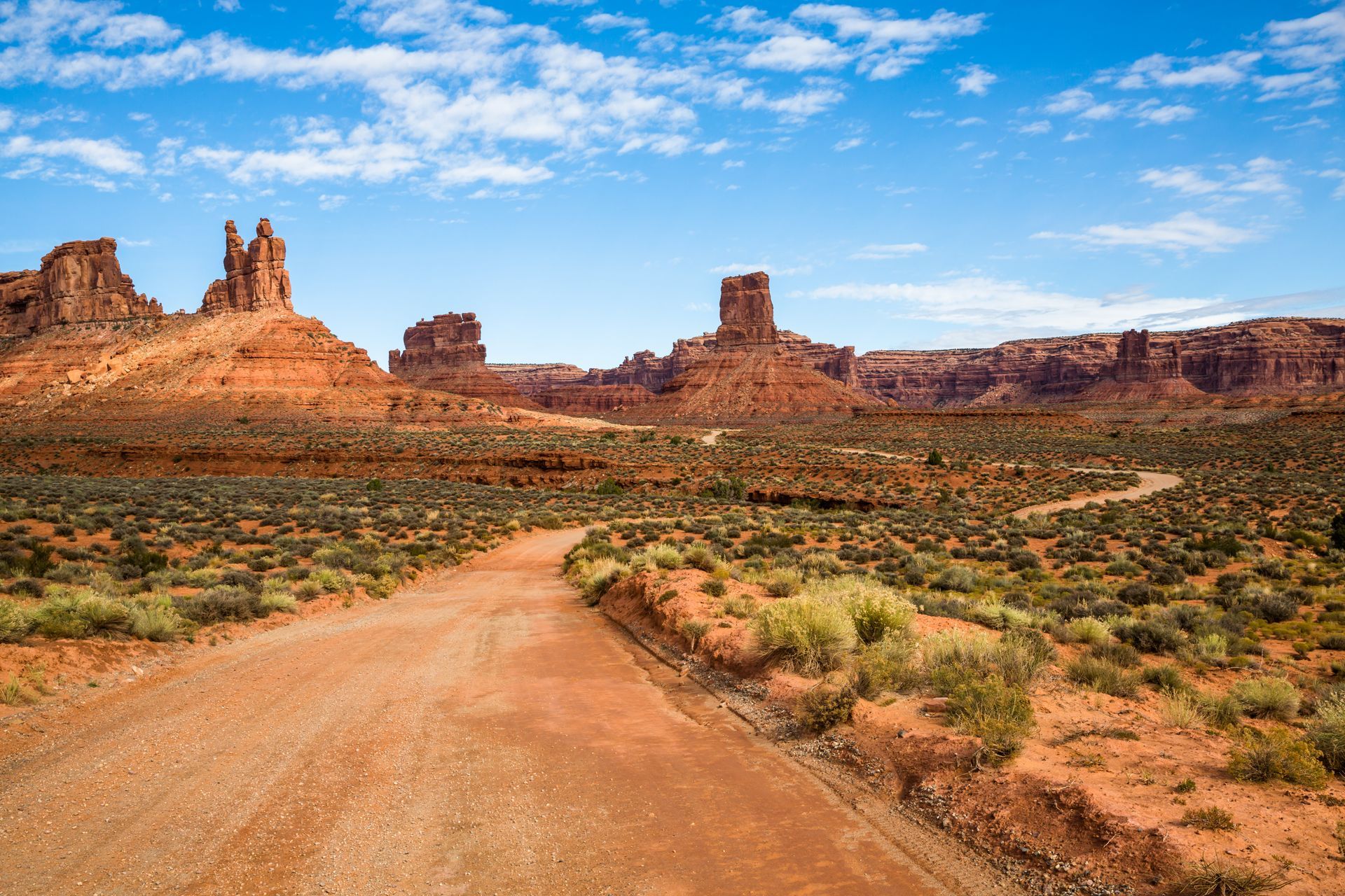 A dirt road leads through a desert landscape with red rock mesas and spires under a bright blue sky with scattered clouds.