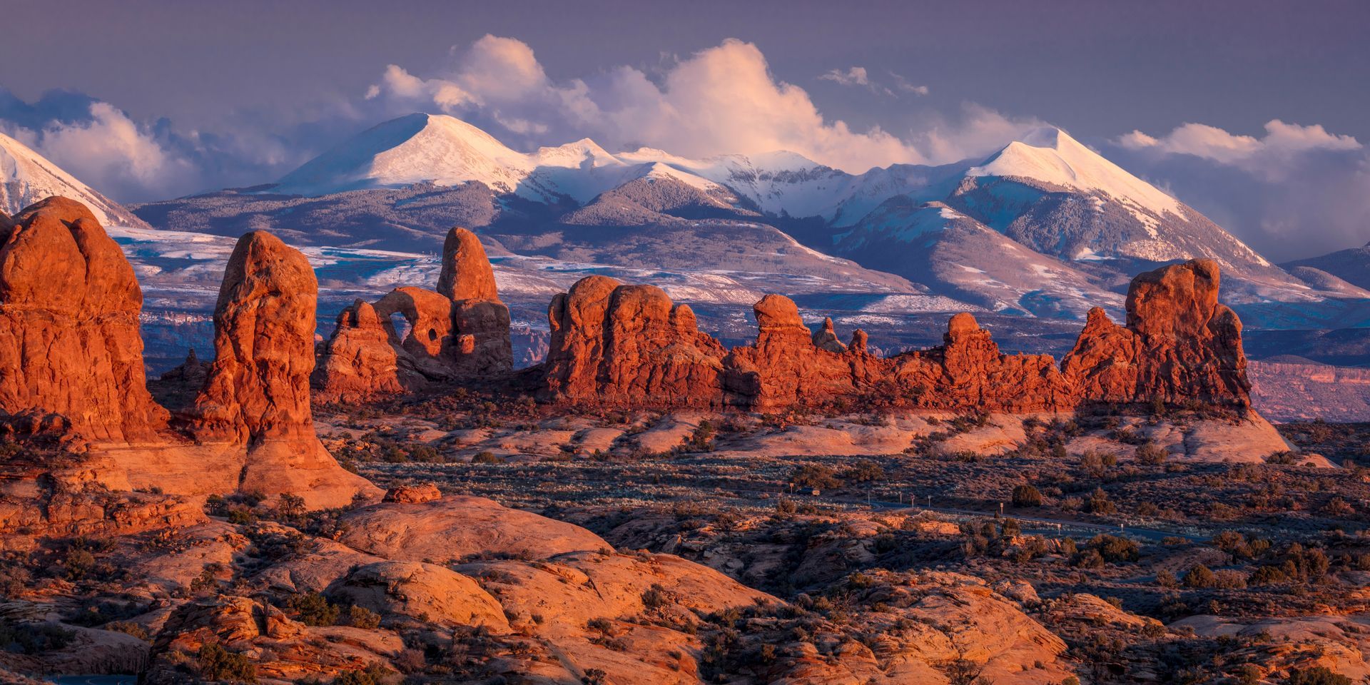 Red rock sandstone formations in Arches National Park under a sunset sky with snow-capped mountains in the background.