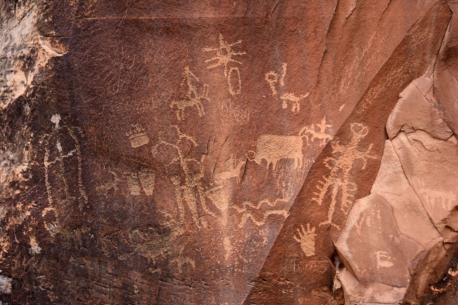 Petroglyphs carved into dark desert rock, featuring human figures, animals, a handprint, and geometric designs.
