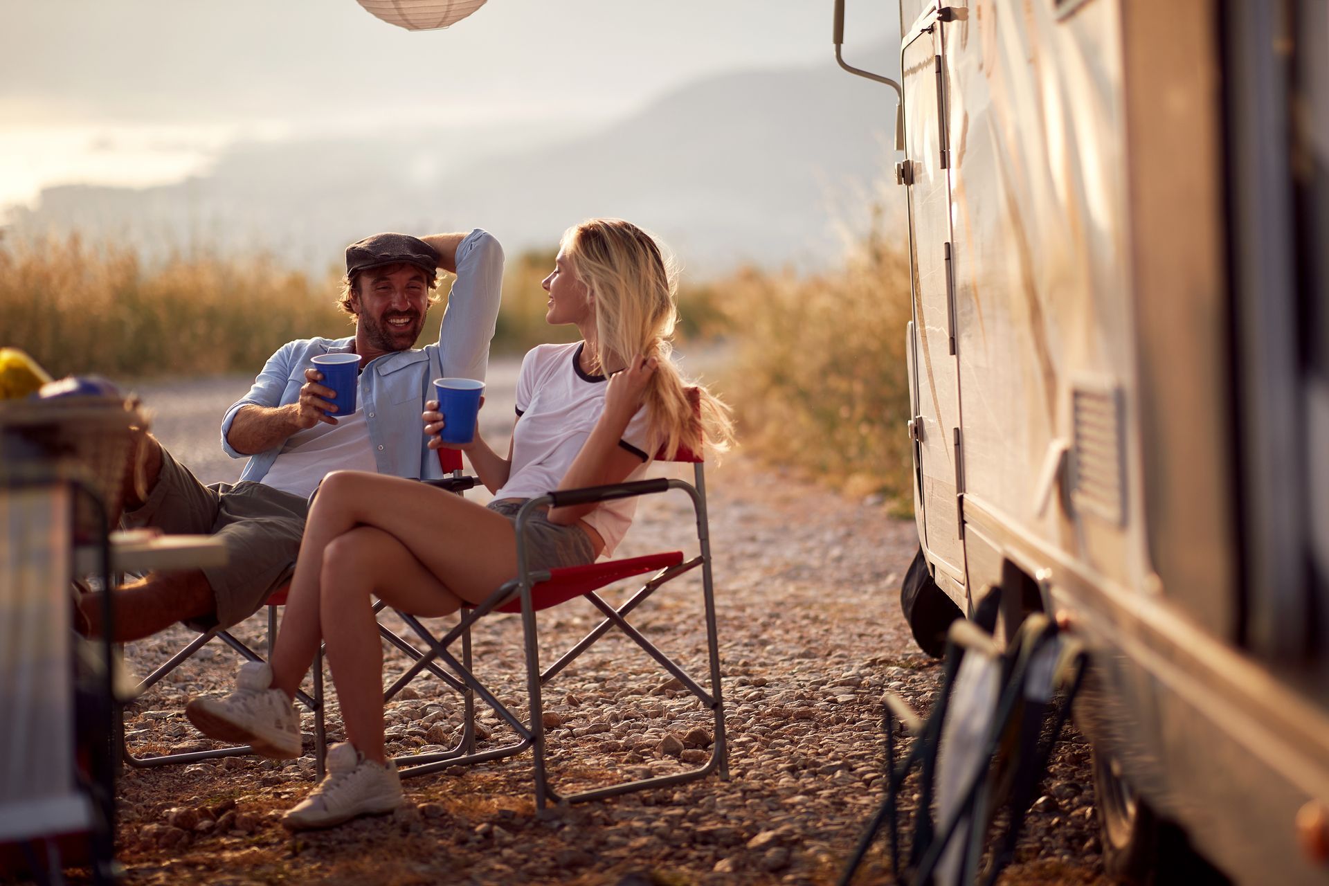 A couple sits in camping chairs outside an RV at sunset, holding blue cups and smiling while looking at each other.