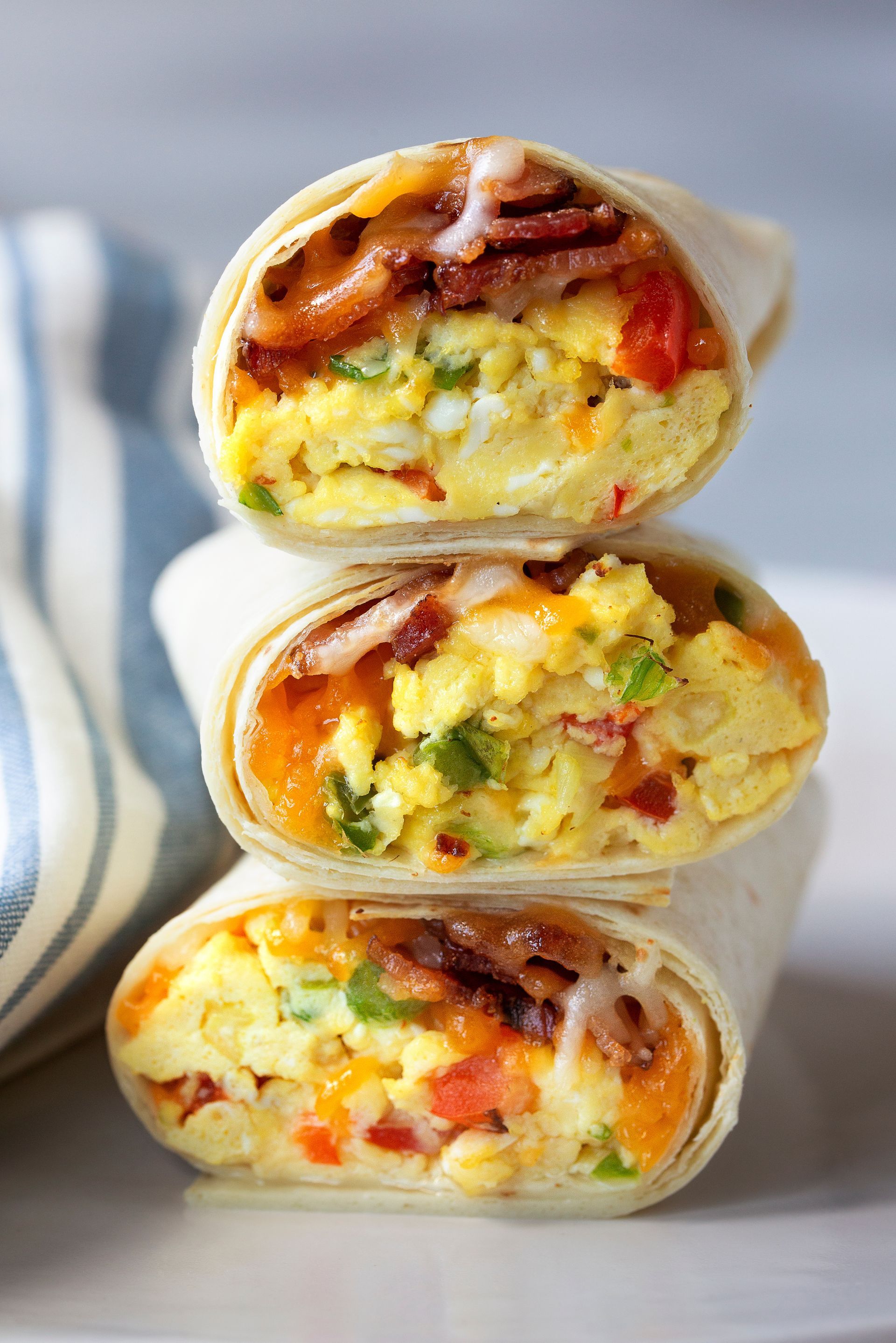 Three breakfast burritos stacked on a white plate, filled with scrambled eggs, bacon, cheese, and red peppers.