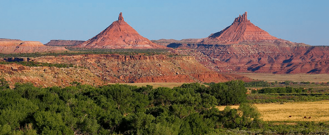 Two prominent red rock peaks rise above a rugged desert landscape with green trees in the foreground under a clear blue sky.