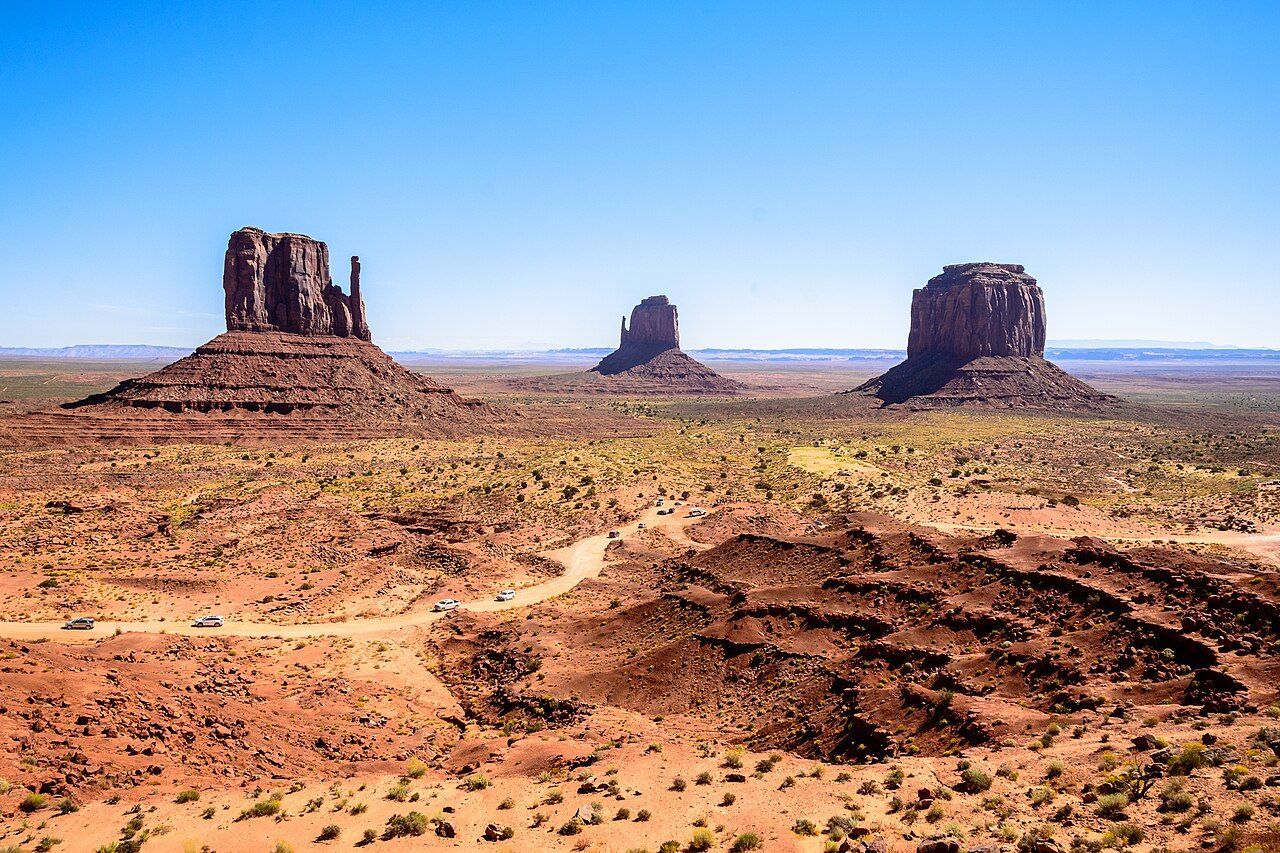 Three iconic sandstone buttes rise above the vast, arid desert landscape of Monument Valley under a clear blue sky.