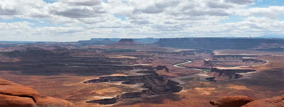 Wide-angle view of a vast, layered desert canyon with red rock formations under a cloudy blue sky.