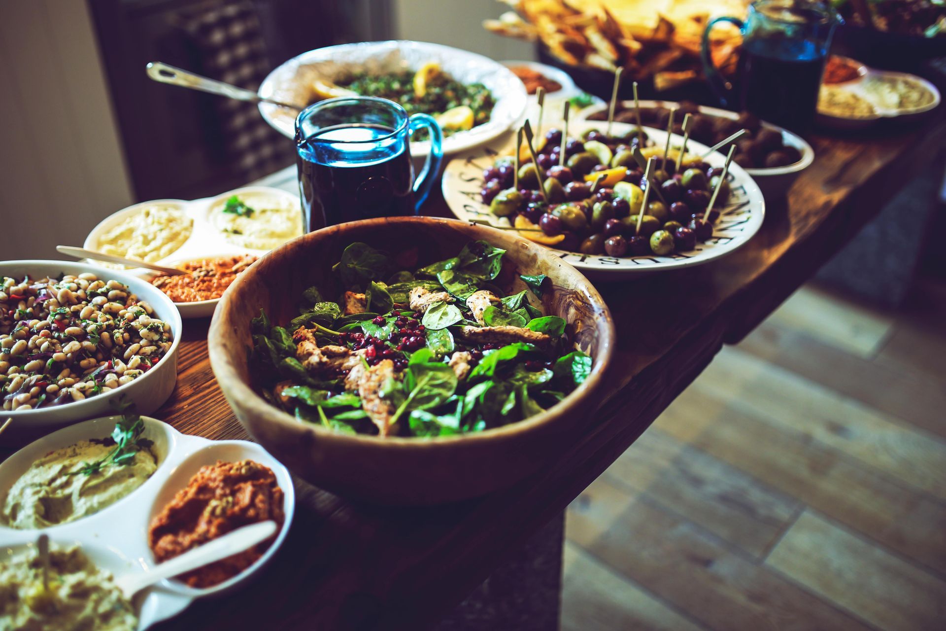 A wooden table laden with a variety of bowls containing salads, dips, olives, and bread in a warm, dimly lit setting.