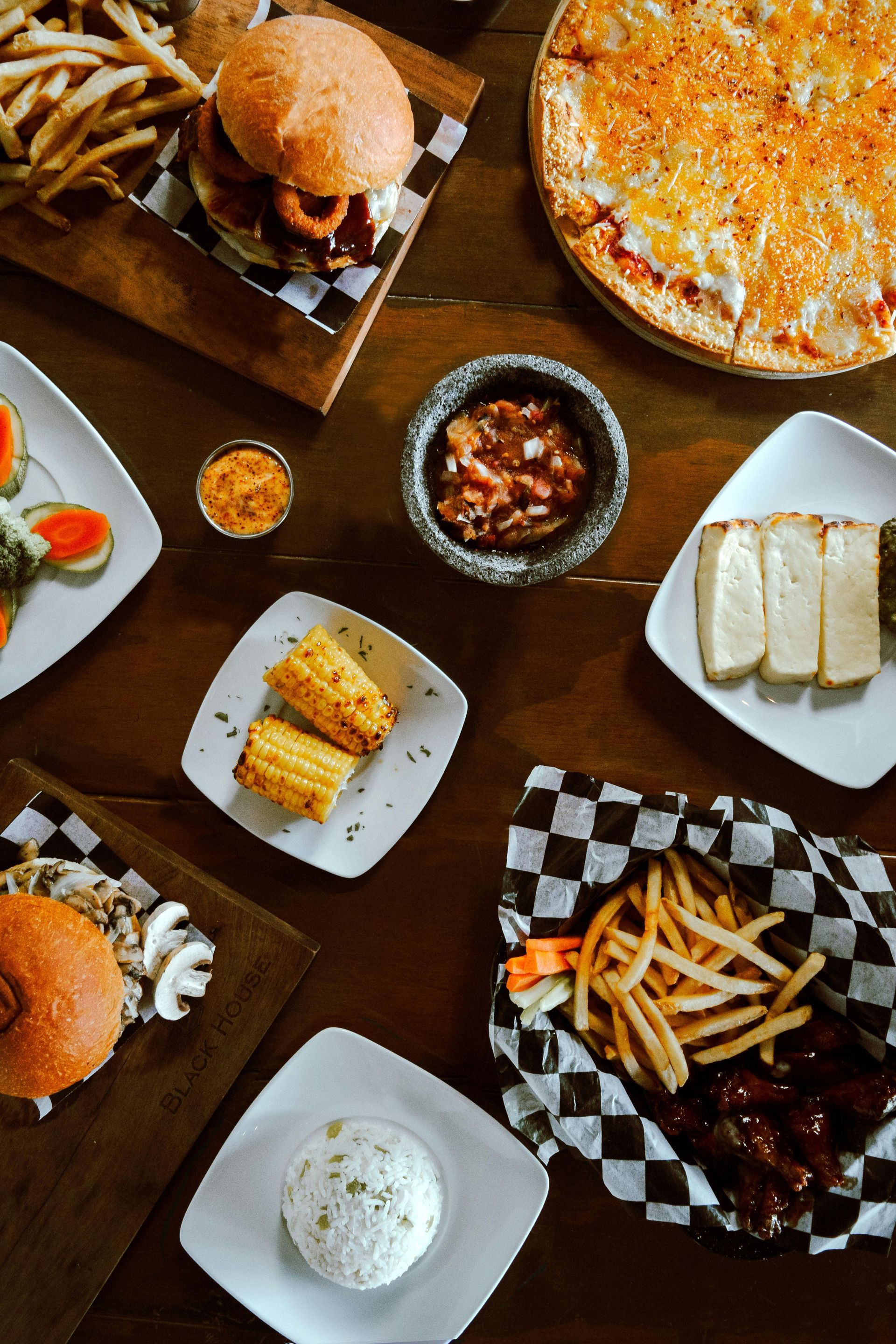 Top-down view of a wooden table spread with burgers, fries, a pizza, corn, dips, and sides on various plates and trays.