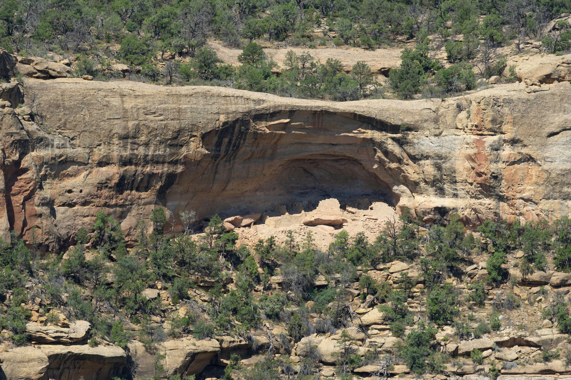 Ancient stone cliff dwellings built into a natural alcove in a rugged, rocky canyon wall with scattered trees.