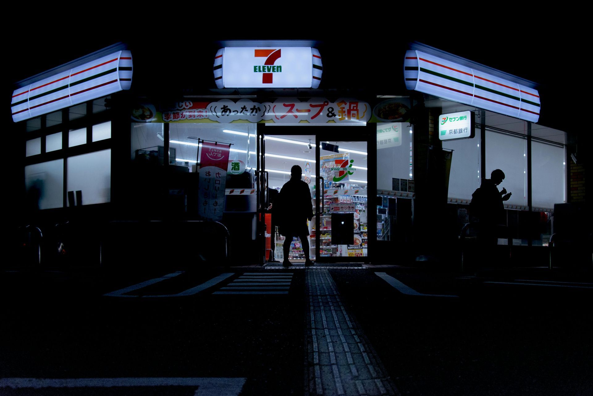 A brightly lit 7-Eleven storefront at night with silhouetted figures standing outside the glass entrance.