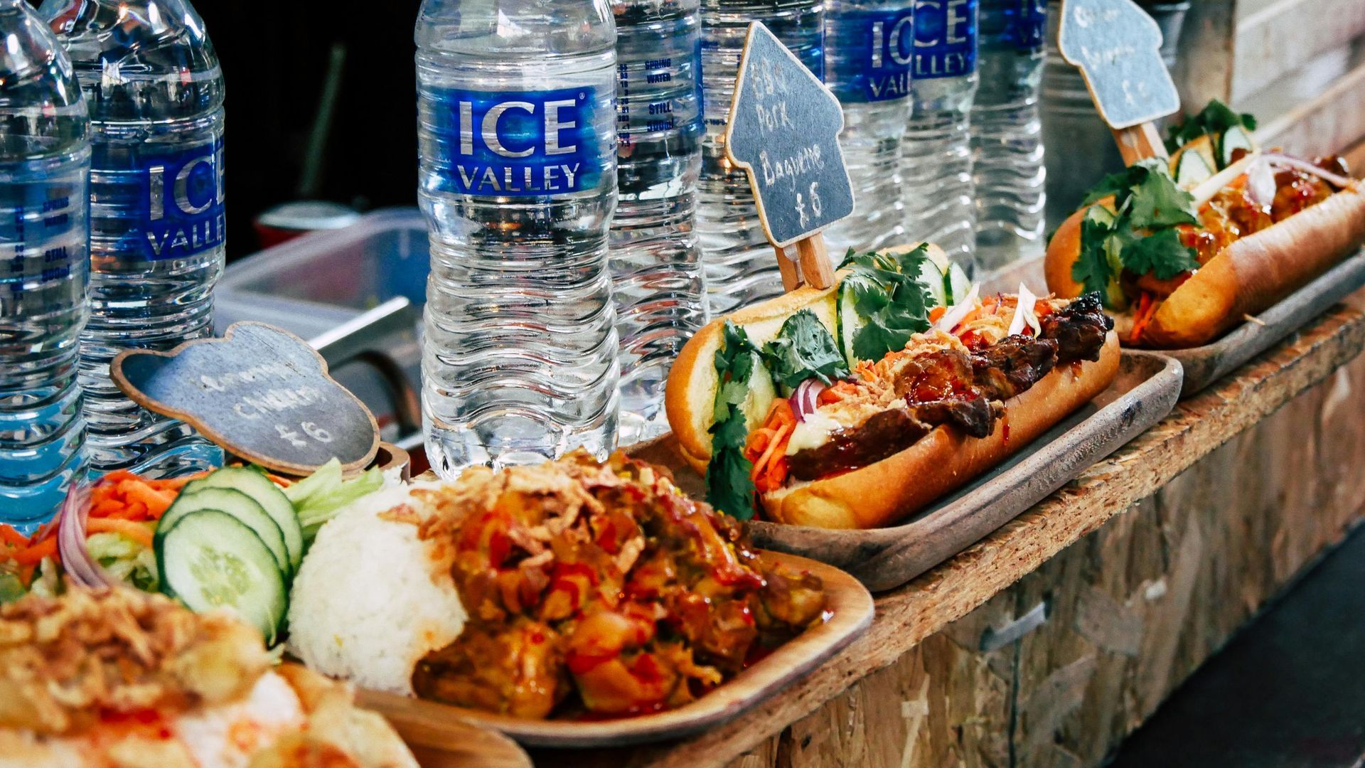 Several bowls of food and hot dogs arranged on a wooden counter with bottles of water in the background.
