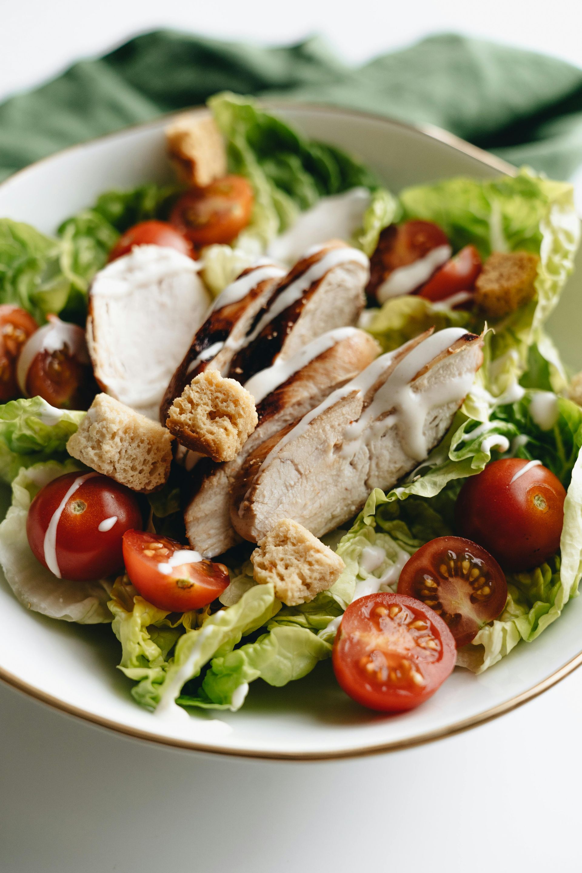 A chicken Caesar salad in a white bowl with cherry tomatoes, croutons, and dressing, against a white background.