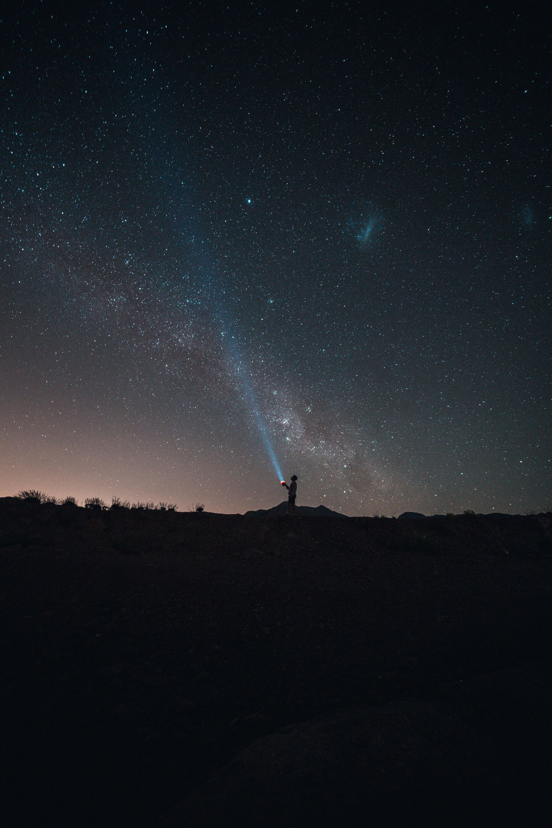 A silhouette of a person standing on a dark horizon, pointing a bright beam of light toward a vast, starry night sky.