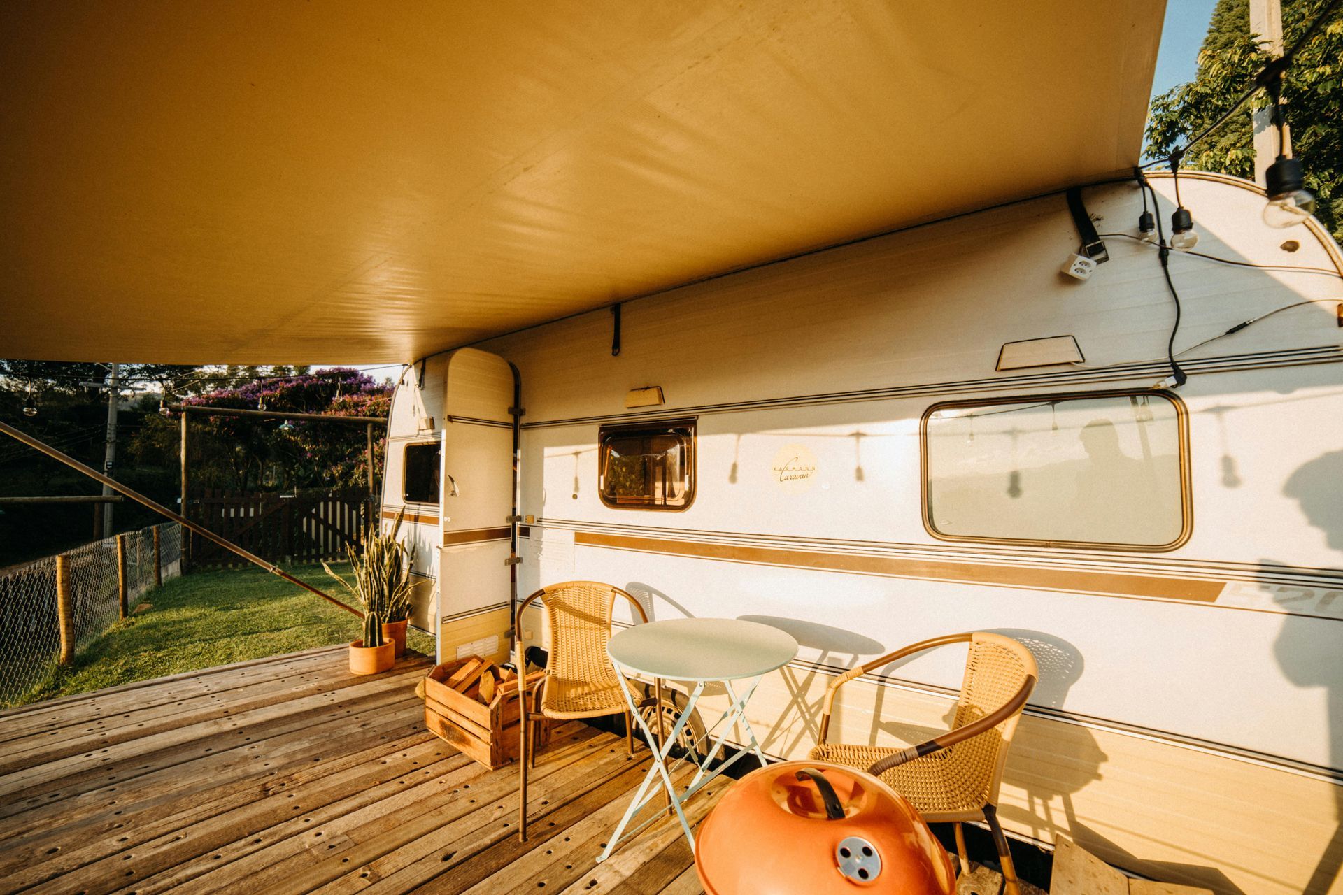 A wooden deck with a small table and two wicker chairs under a camper awning, next to a white vintage travel trailer.