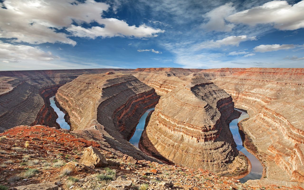 A river meanders in tight, deep loops through a vast, arid canyon with tiered, rocky cliffs under a blue sky.