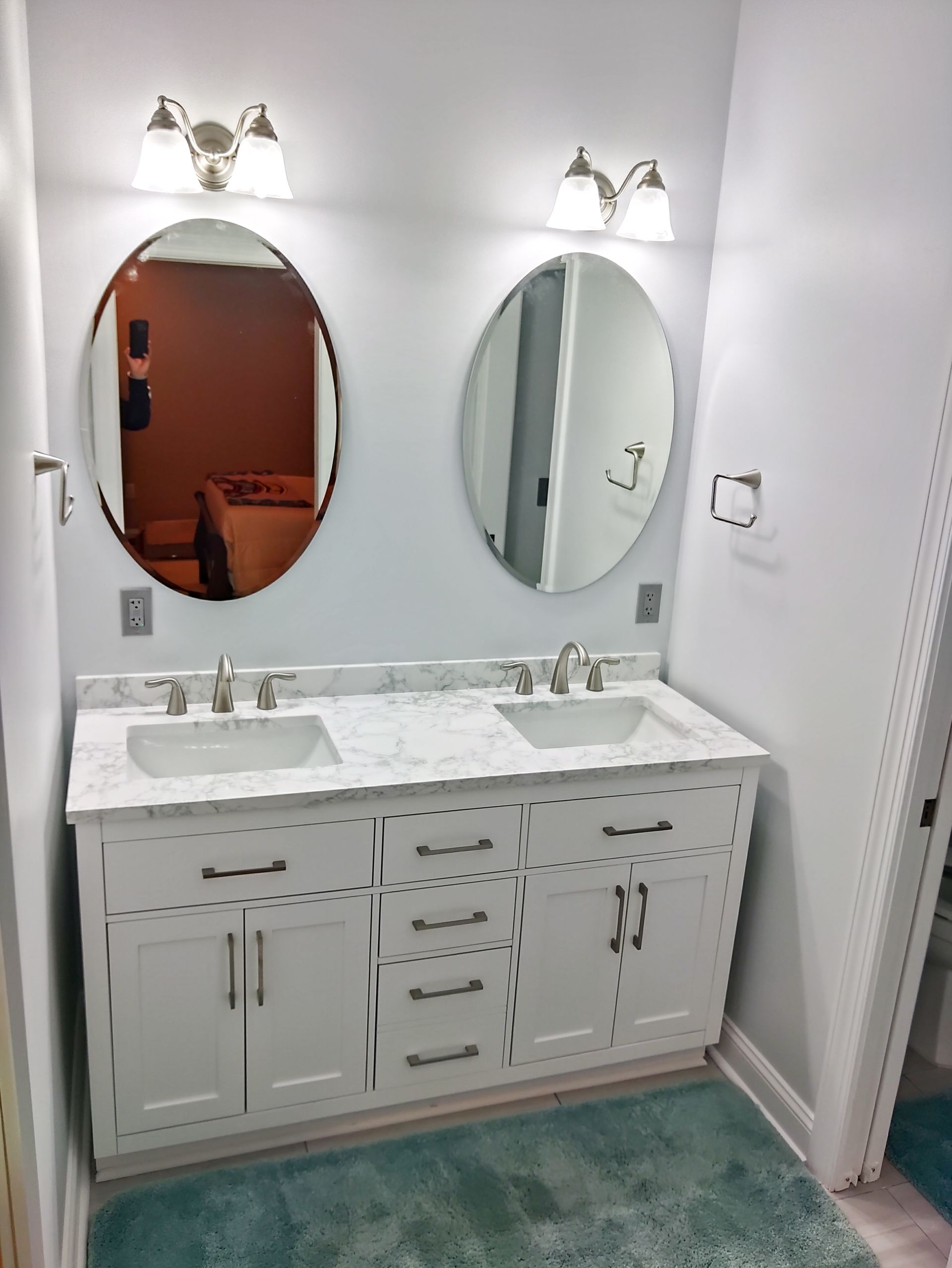 White bathroom vanity with double sinks, oval mirrors, and wall sconces over a teal rug.