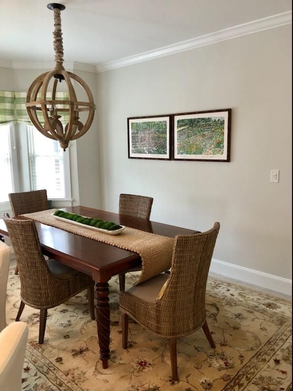 Dining room with wicker chairs, wooden table, patterned rug, and two framed artworks on a light wall.