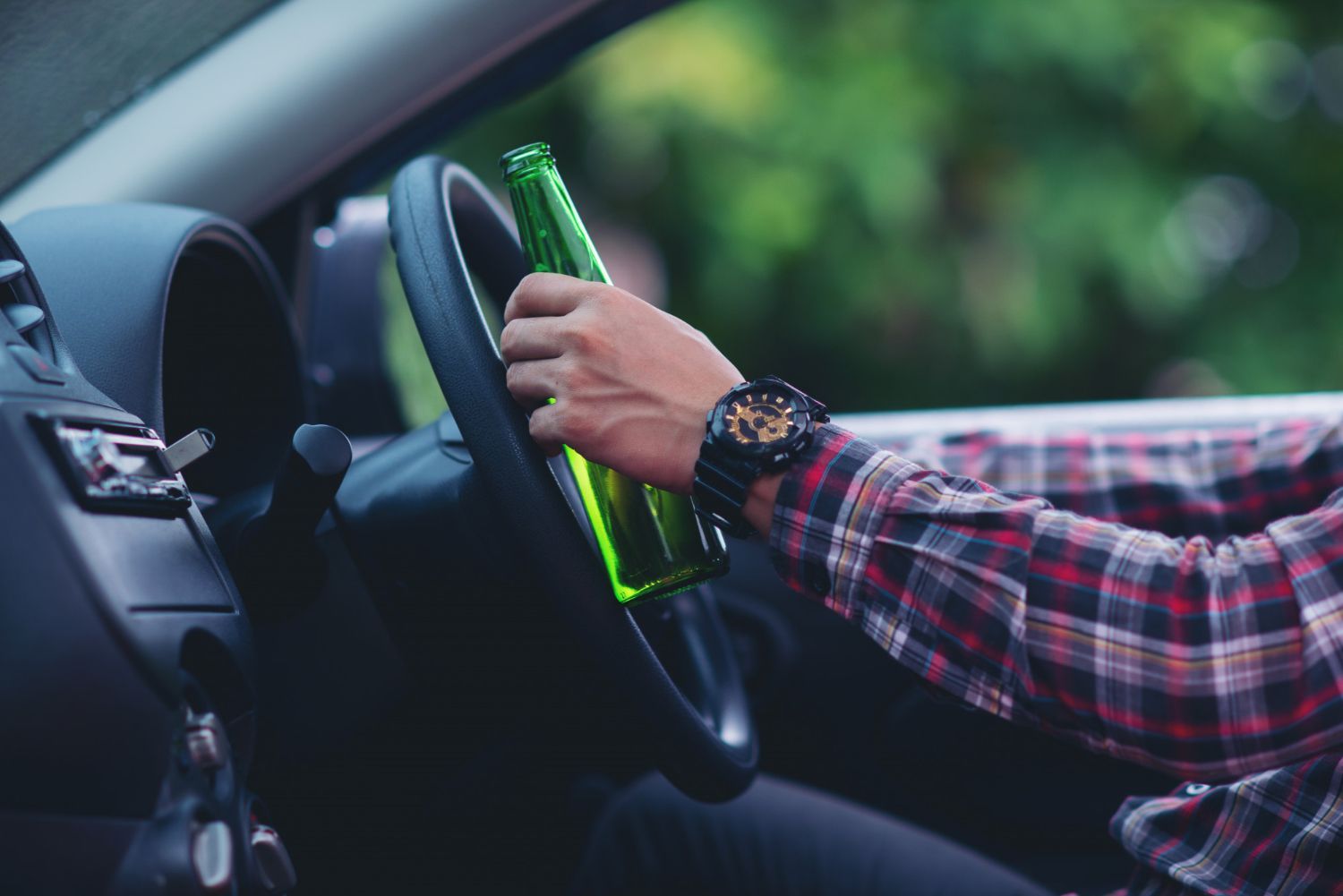 Person in a car holding a green beer bottle while gripping the steering wheel.