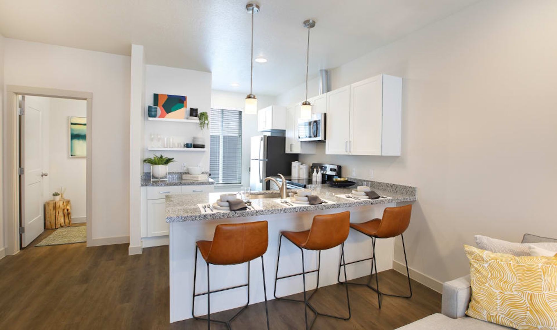 Kitchen with granite countertop, bar stools, and white cabinets. Brown flooring, and a glimpse of a bedroom.