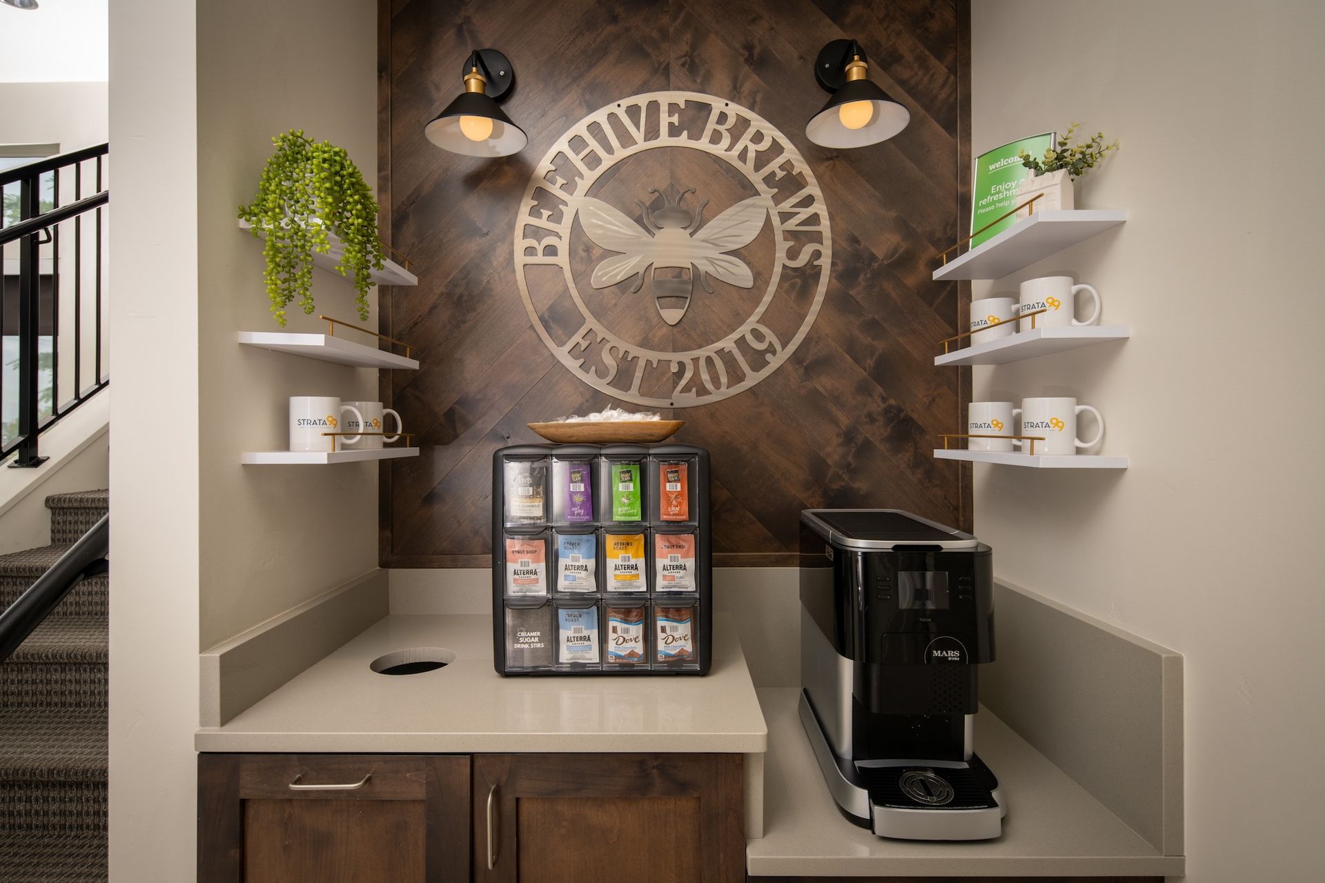 Coffee station with tea, coffee maker, and decorative bee logo on wood paneling.