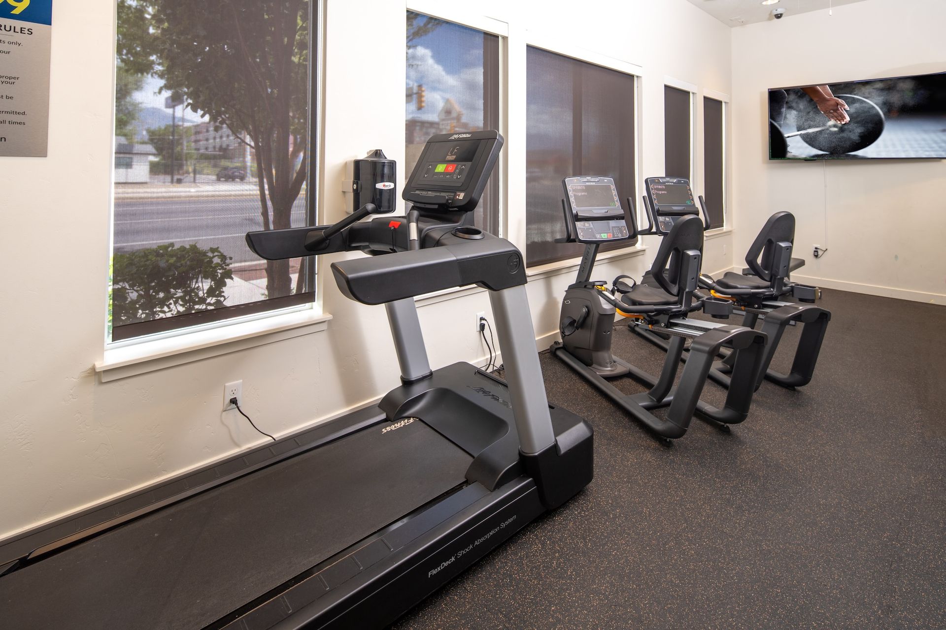 Gym with treadmill and recumbent bikes, next to windows and TV.