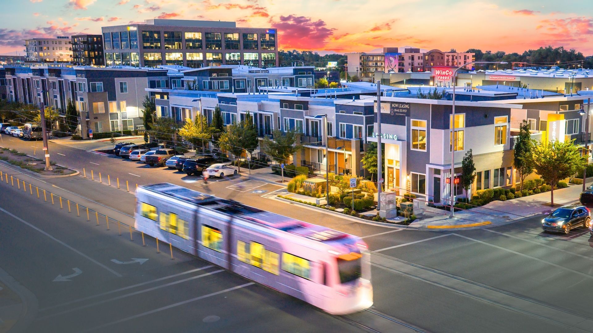 A light rail train travels through a modern cityscape at dusk; residential buildings and an office building in the background.