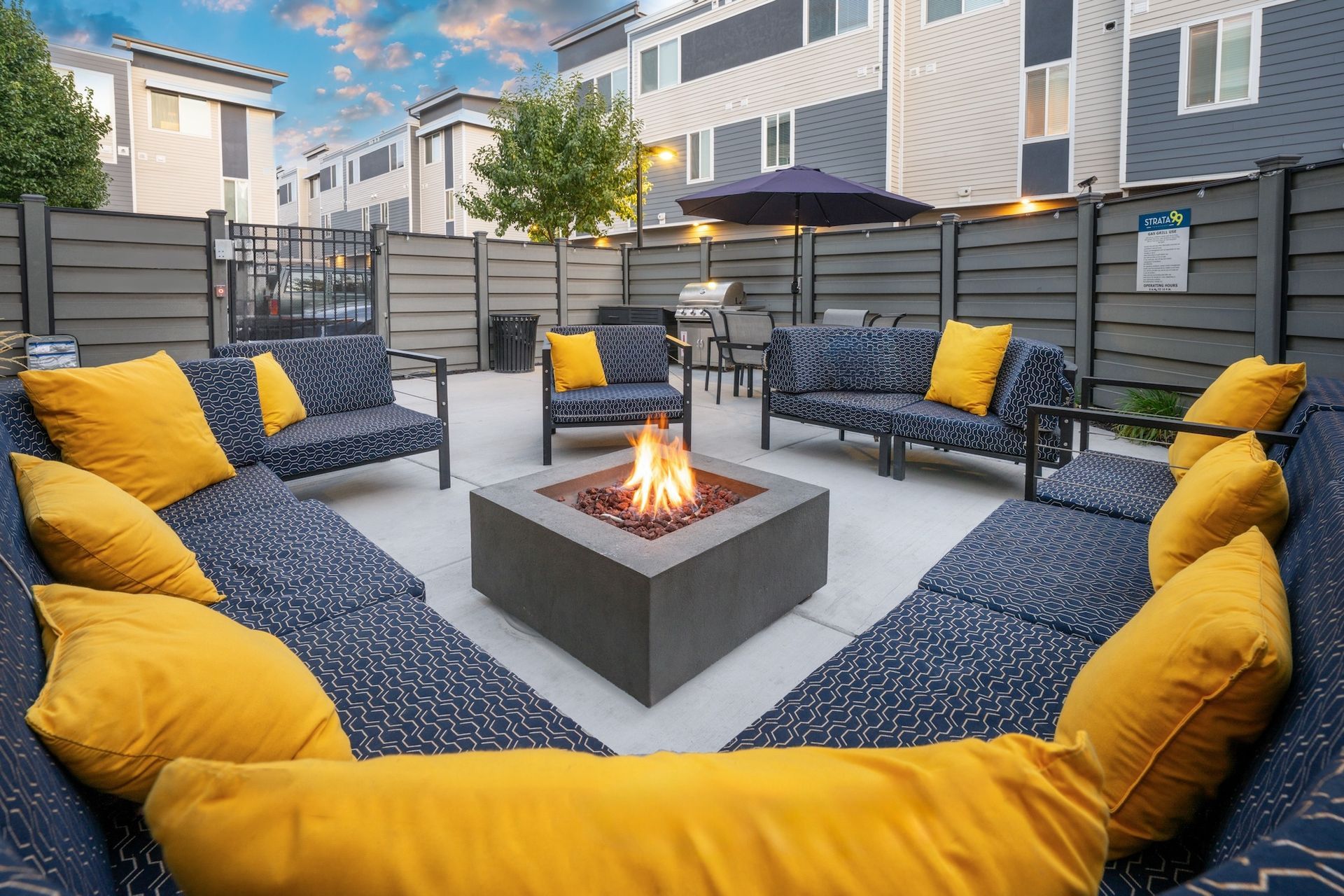 Outdoor lounge with fire pit and seating, blue and yellow cushions, fenced area, buildings in background.
