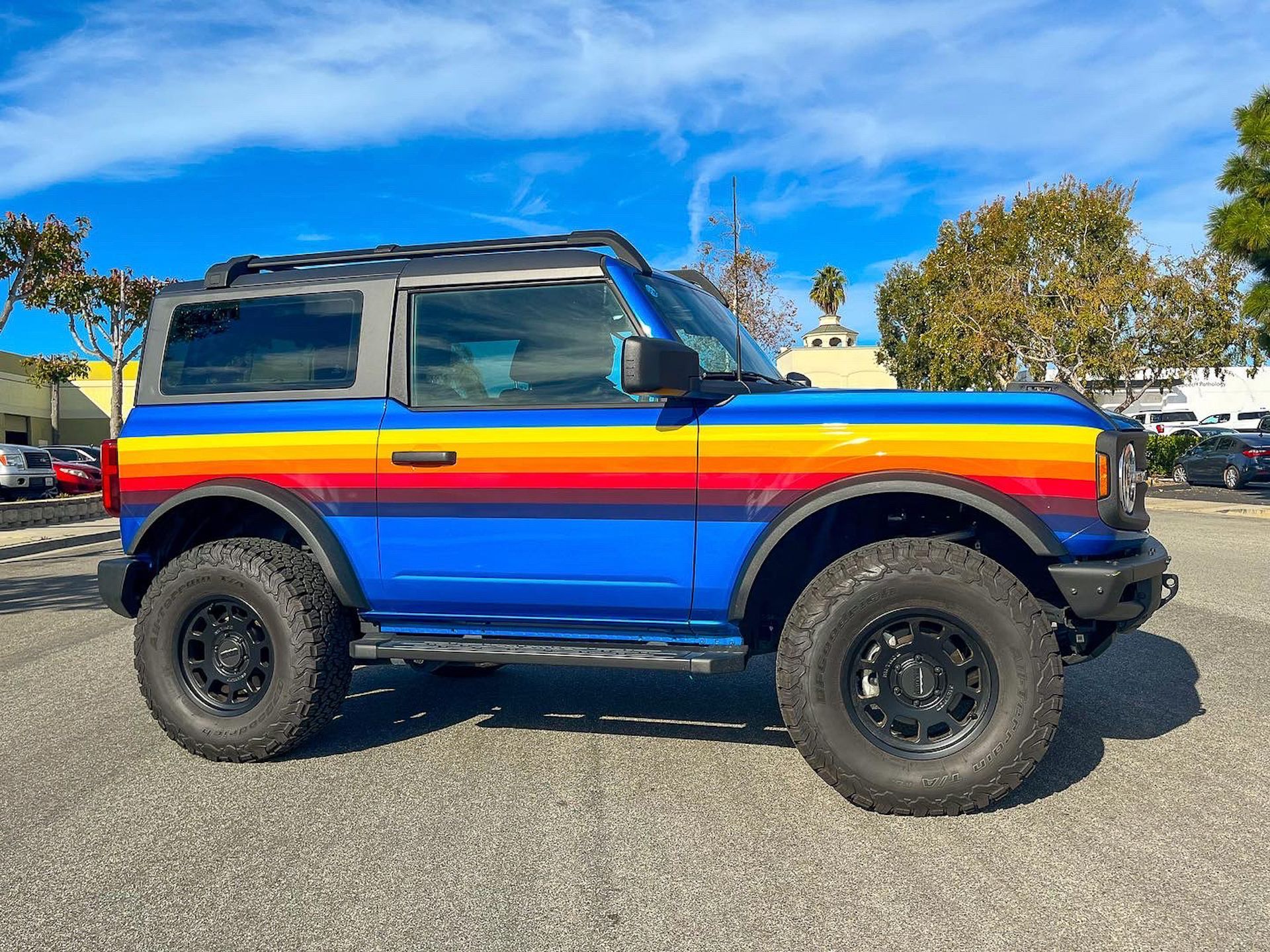 Blue Ford Bronco with rainbow stripe, black wheels, roof rack, parked on asphalt under a blue sky.