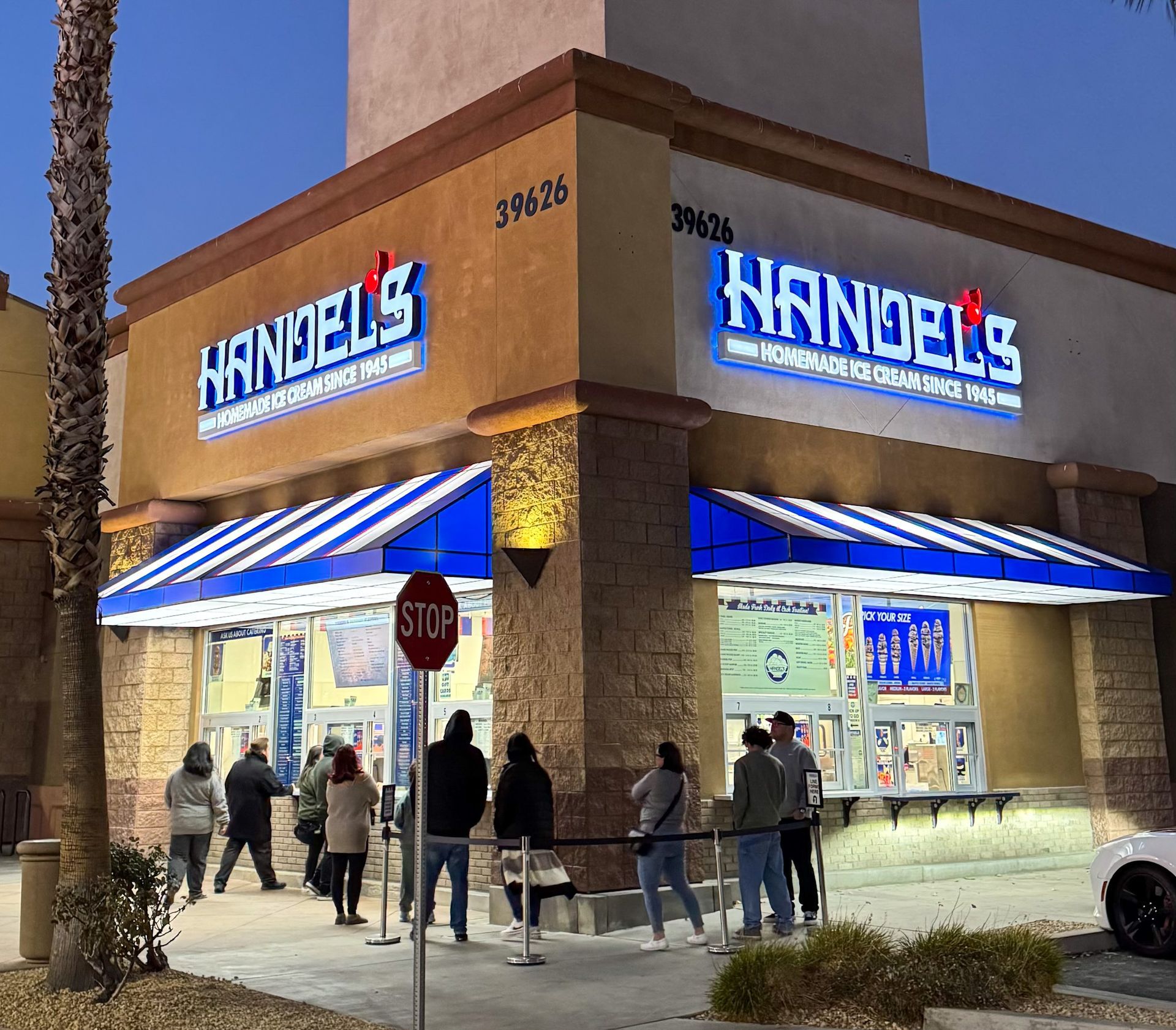 Handel's ice cream shop at dusk with people lined up. Blue and white awning, illuminated sign, Stop sign.