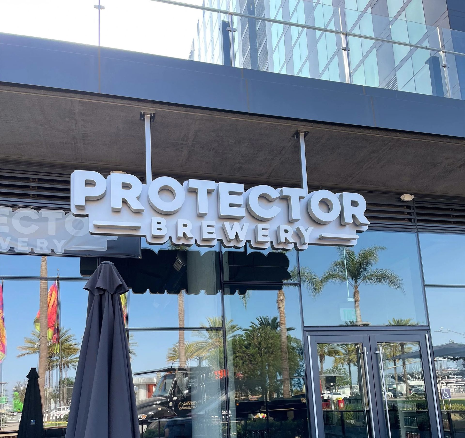 Protector Brewery storefront with white letters. Building has glass windows, a dark umbrella, and palm trees.
