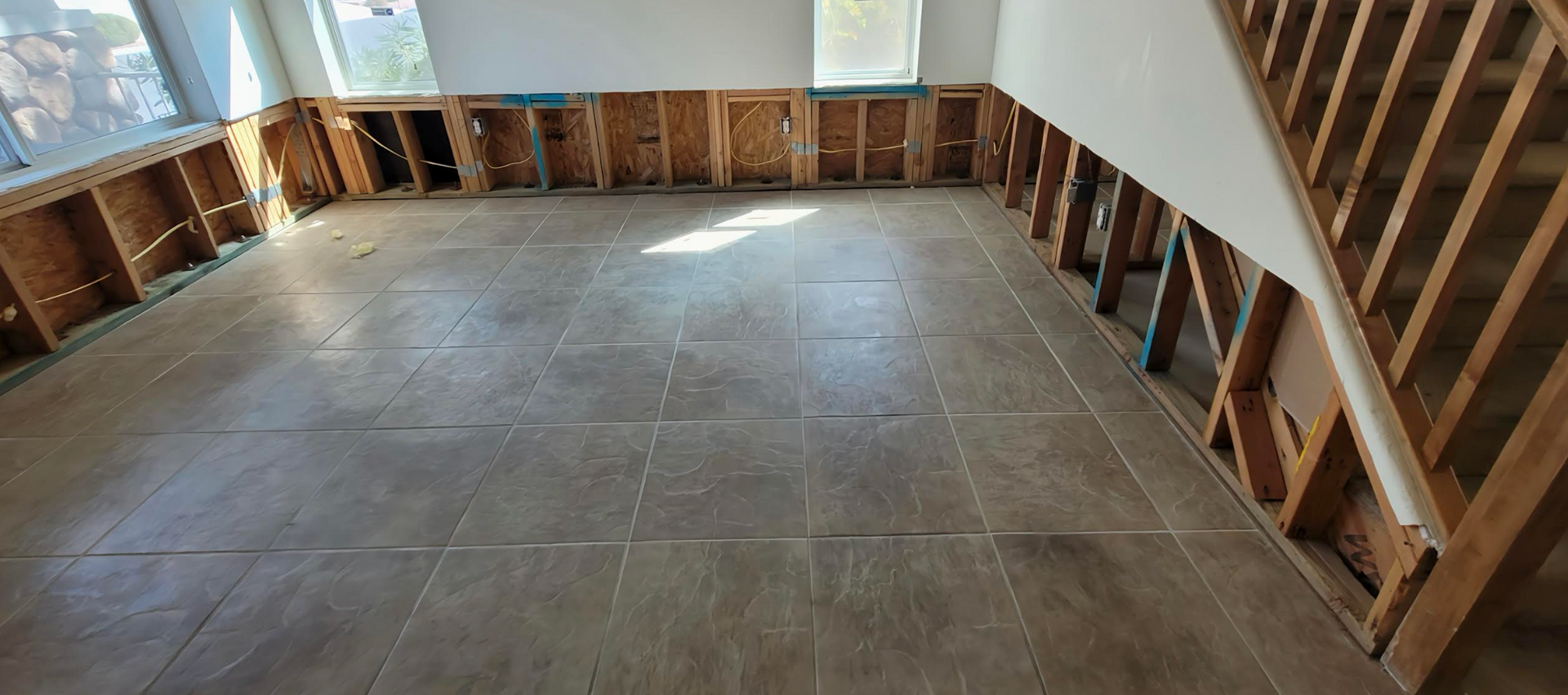 Interior room undergoing renovation, exposed wall studs, flooring, staircase, windows, sunlight.