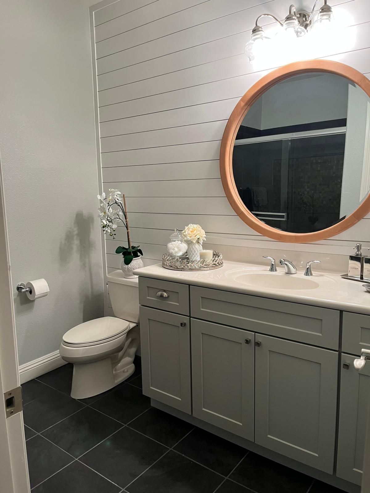 A bathroom featuring a gray vanity, white countertop, round mirror, and a shiplap accent wall. Black tile floor and white toilet.