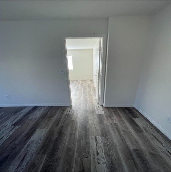 An empty room with gray wood flooring, white walls, and a doorway leading to another room.