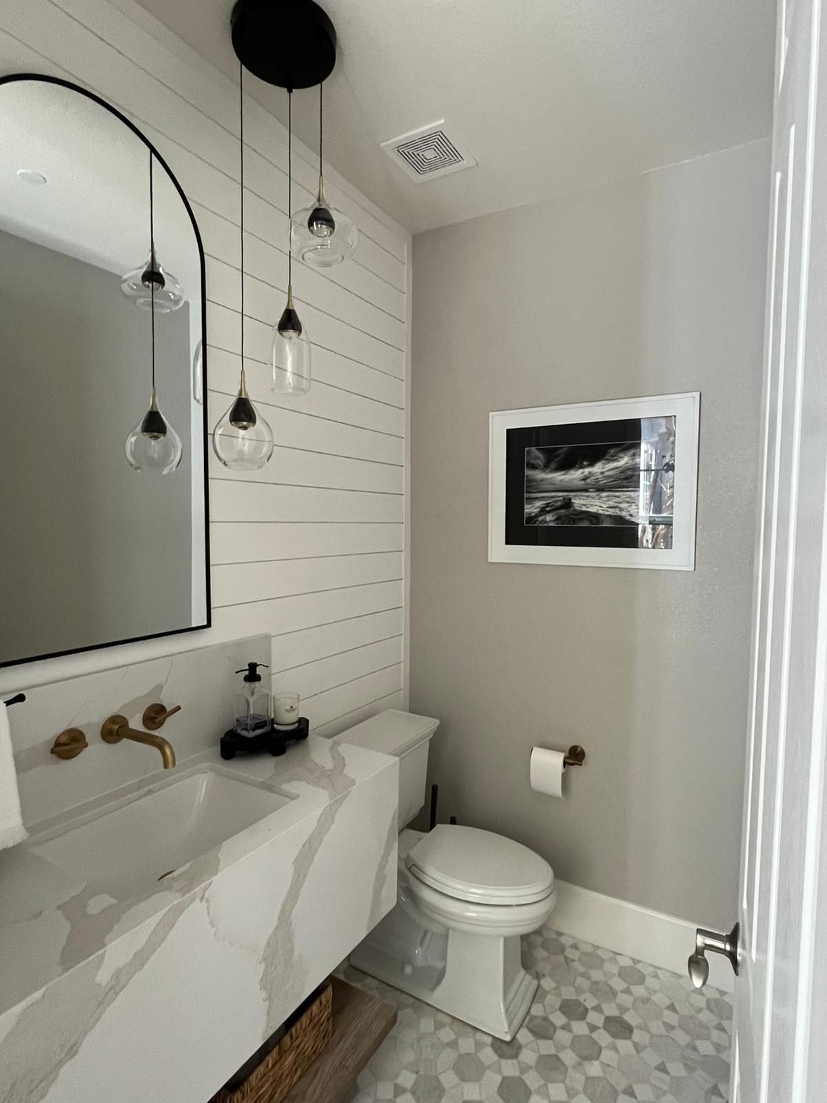 A modern bathroom with a white marble countertop, a shiplap wall, and an arched mirror. There are pendant lights and a framed ultrasound image.