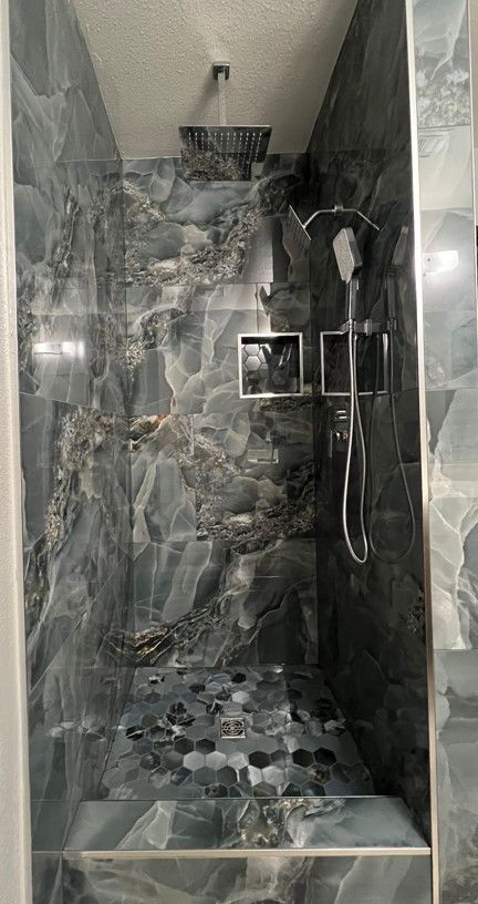 A walk-in shower with dark blue and gray marbled tile. The floor is made of dark, smooth stones. Showerhead and handheld sprayer visible.