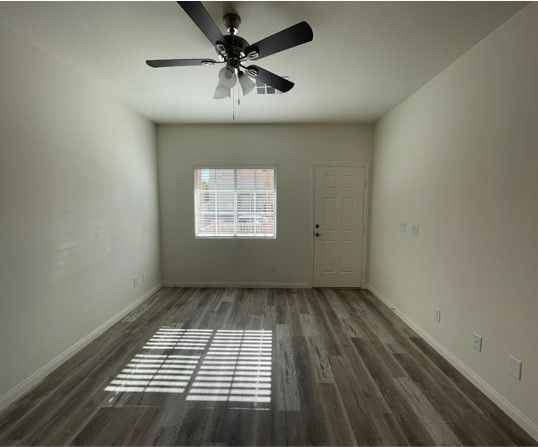 An empty room with wood-look flooring, light walls, a ceiling fan, and a window with sunlight casting shadows.