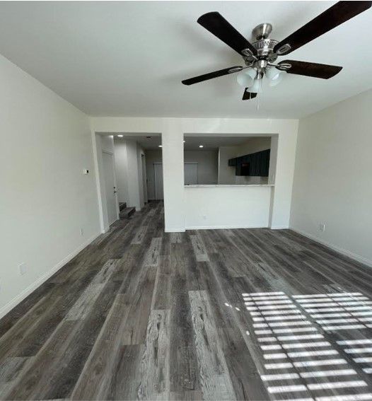 Empty living room with dark wood-look flooring, white walls, and a ceiling fan. Sunlight streams in from the right.