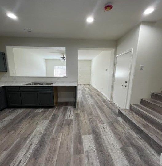 Interior view of a kitchen with dark gray cabinets, open to a hallway with wood-look flooring. White walls and a staircase are visible.